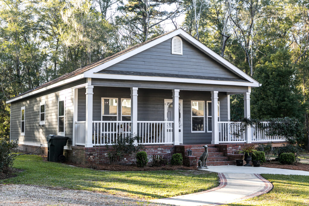 Small blue gray mobile home with a front and side porch. It has a white porch railing out in the country on land shared by a larger siding house.