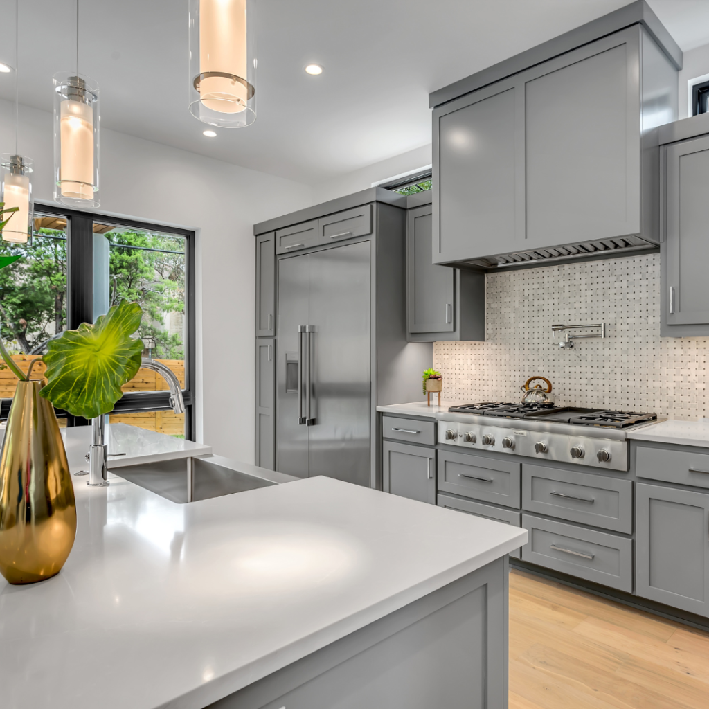 Kitchen with gray cabinets and gray countertops, stainless steel refrigerator, 3 cylindrical pendant lights clear and silver, bright natural light